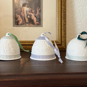 May include: Three white ceramic bell ornaments with embossed designs and coloured ribbons. The bells are dated 1992, 1993, and undated. The background includes a framed picture and a dark wooden surface.