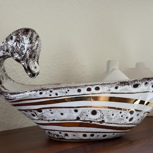 May include: A ceramic swan-shaped bowl featuring a white base with a speckled dark brown glaze and horizontal metallic gold stripes. The elongated neck of the swan curves gracefully to form one end of the vessel, while the body is elongated.