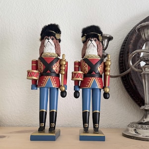 Pode incluir: Duas figuras de quebra-nozes idênticas em forma de cão com pelo castanho e branco, usando chapéus pretos altos e felpudos, casacos vermelhos com tiras pretas cruzadas, calças azuis e botas pretas. Cada figura segura um pequeno tambor.