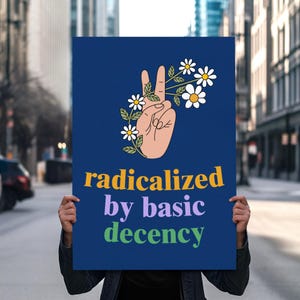 Pode incluir: Um cartaz azul marinho com uma mão fazendo o sinal de paz, adornado com flores de margarida. O texto "radicalized by basic decency" está em amarelo, roxo e verde. O cartaz é segurado em frente a uma rua da cidade.