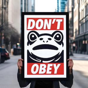 Puede incluir: Un póster enmarcado en negro con las palabras "DON'T OBEY" en letras blancas sobre un fondo rojo. El póster presenta una ilustración estilizada de una rana en blanco y negro. La imagen se sostiene frente a una calle de la ciudad borrosa.