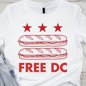 May include: White t-shirt with a red graphic. The graphic features three red stars above two sandwich illustrations. Below the sandwiches, the text "FREE DC" is printed in red. The shirt is made of a soft material.