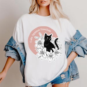 May include: White t-shirt with a graphic featuring a black cat surrounded by white lilies, set against a pink circle. The text "Cats Against Fascism" is arched around the circle. The shirt is worn with a denim jacket.