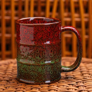 May include: A ceramic mug with a textured surface and a handle. The mug features a gradient of colors, starting with red at the top, transitioning to green in the middle, and ending with brown at the bottom. The mug is sitting on a woven surface.