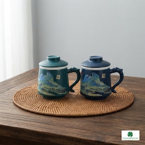 May include: Two ceramic mugs with lids and handles, one in teal and one in dark blue. Each mug features a painted landscape scene of mountains and water in shades of blue, green, and gold, with a small square stamp detail on the side.