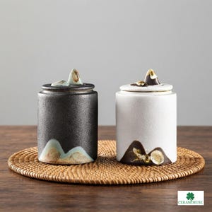 Könnte beinhalten: Zwei zylindrische Keramikdosen, eine schwarz und eine weiß, mit strukturierter Oberfläche und einem Deckel, der mit einem kleinen bergförmigen Griff versehen ist. Der Boden jeder Dose ist mit einem Bergmotiv in erdigen Farbtönen bemalt.