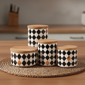 Puede incluir: Un juego de cuatro botes de cocina de cerámica con tapas de madera. Los botes presentan un patrón de diamantes en blanco y negro con un diamante de color cobre. Los botes están colocados sobre un salvamanteles tejido.