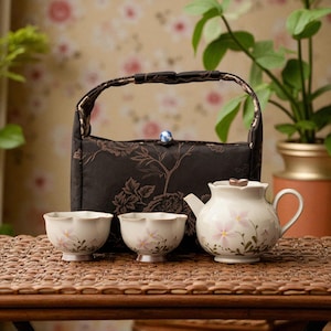 Op de afbeelding: Een theeservies met een witte theepot en twee bijpassende kopjes, elk versierd met roze bloemmotieven. Een zwarte draagtas met een bloemmotief en een handvat is ook inbegrepen. De set wordt gepresenteerd op een geweven oppervlak.