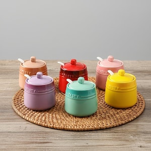 May include: Six colorful ceramic sugar bowls with lids and small spoons. The bowls are in shades of pink, yellow, green, purple, red, and peach. Each bowl has the word "TASTY" embossed on the side. The bowls are arranged on a woven mat.