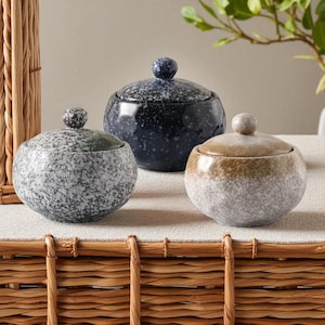 May include: Three decorative lidded jars in a speckled stone finish. The jars are round with a small knob on top of the lid. The colors are gray, navy blue, and tan. The jars are sitting on a woven surface.