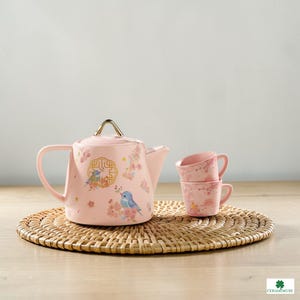 Pode incluir: Uma chaleira de cerâmica rosa e duas chávenas a condizer com um design floral e de pássaros. A chaleira tem uma pega dourada e uma tampa. As chávenas estão empilhadas. O conjunto está sobre uma esteira tecida.