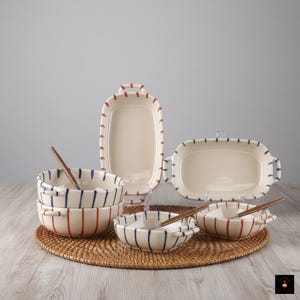 Op de afbeelding: Een verzameling crèmekleurige keramische serveerschalen en kommen met rood en blauw gestreepte accenten. De set bevat rechthoekige schalen met handvatten, kommen en houten lepels, gerangschikt op een geweven mat.