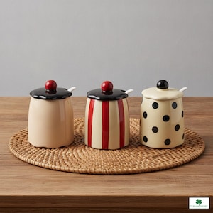 Op de afbeelding: Drie keramische potten met deksels en kleine lepels. De eerste is effen beige met een zwart deksel en rode knop. De tweede heeft rode en beige verticale strepen, een zwart deksel en een rode knop. De derde is beige met zwarte stippen en een zwart deksel.