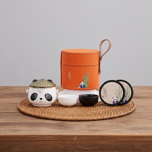 May include: A ceramic tea set featuring a panda theme. The set includes a teapot with a panda face, two small cups, a carrying case, and two coasters. The case is orange with a panda and bamboo design. The set is on a woven mat.