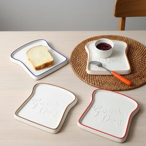 Puede incluir: Platos blancos de cerámica con forma de rebanada de pan, con bordes de colores. Uno contiene una rebanada de pan, otro un cuenco pequeño con mermelada y un tenedor. Los platos tienen la inscripción "Good Day".
