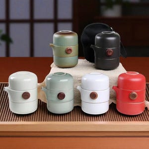 May include: A collection of ceramic tea sets in various colors, including green, white, and red. Each set has a spout and a wooden button detail. A black carrying case is also included.