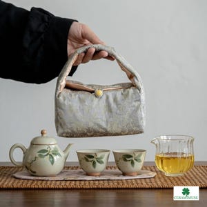 Puede incluir: Un portatetera de tela acolchada gris claro con estampado floral y un botón amarillo. A su lado hay una tetera de cerámica y dos tazas a juego con diseños de hojas verdes, además de una jarra de cristal con té.