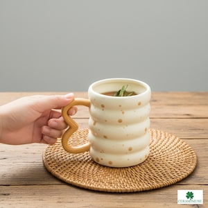 Può includere: Tazza in ceramica color crema con un design a coste unico e un manico color senape. La tazza ha macchie marroni e contiene una bevanda con erbe. È appoggiata su un sottobicchiere intrecciato.