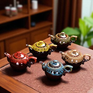 May include: A collection of five ceramic teapots in various colors, including red, yellow, brown, green, and blue. Each teapot has a unique design and a small handle. The teapots are displayed on a wooden table.