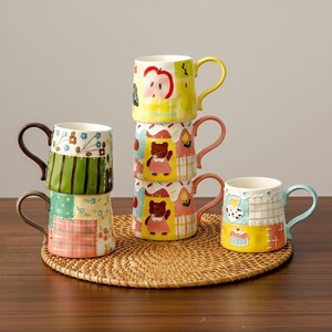 Könnte beinhalten: Ein Stapel von sechs Keramikbechern mit bunten Patchwork-Designs. Die Becher zeigen verschiedene Illustrationen, darunter einen Apfel, einen Bären und einen Cupcake. Jeder Becher hat einen unterschiedlich farbigen Henkel und steht auf einer gewebten Matte.