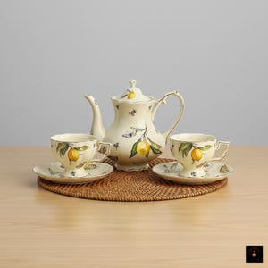 Puede incluir: Juego de té color crema con tetera, dos tazas y platillos. El juego está decorado con limones amarillos, hojas verdes e ilustraciones de mariposas. El juego está colocado sobre un salvamanteles redondo y tejido.