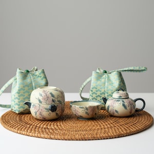 May include: A ceramic tea set with floral designs, including a teapot, teacup, and a tea container, sits on a woven mat. Two teal fabric pouches with green accents are also present. The set is ideal for tea lovers.