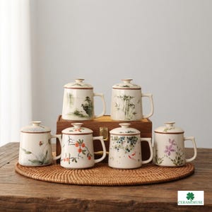 May include: Six off-white ceramic mugs with lids, each featuring unique hand-painted botanical or landscape designs including bamboo, flowers, and fruit. The mugs have brown rim accents and handles, arranged in two rows on a woven circular mat.