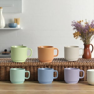 May include: A collection of six ceramic mugs in various pastel colors, including green, orange, cream, teal, blue, and lavender. Each mug has a ribbed design and a handle. The mugs are arranged on a woven surface.