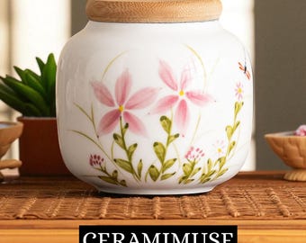 May include: White ceramic jar with a wooden lid, adorned with pink floral designs and green leaves. The jar has a rounded shape and is sitting on a woven surface. The text "CERAMIMUSE" is displayed at the bottom.