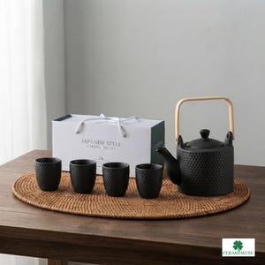 Könnte beinhalten: Ein matt-schwarzes Keramik-Teeservice bestehend aus einer Teekanne mit hellem Holzgriff und vier passenden Tassen, alle mit einer strukturierten Wabenoptik. Das Set wird mit seiner weißen Verpackungsschachtel mit der Aufschrift JAPANESE STYLE CERAMIC TEA SET gezeigt.
