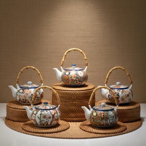 Puede incluir: Cinco teteras de cerámica decorativas con asas de bambú. Las teteras tienen cuerpos blancos con diseños florales y geométricos en tonos naranja, azul y verde. Se encuentran sobre esteras y soportes de ratán tejido.