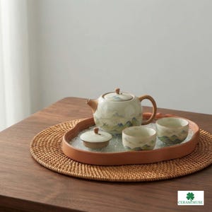 Pode incluir: Um conjunto de chá em cerâmica composto por um bule com tampa, duas chávenas pequenas e um pequeno recipiente com tampa, decorados com uma paisagem montanhosa pintada em tons suaves de azul e verde, dispostos num tabuleiro cor de terracota.