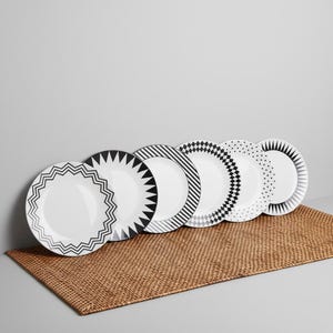 May include: A set of six white ceramic plates with black and gray geometric patterns. Each plate features a unique design, including zigzags, triangles, stripes, and diamonds. The plates are arranged on a woven brown placemat.