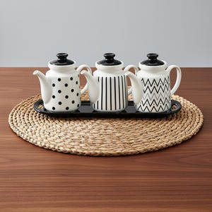 May include: Three white ceramic teapots with black lids and handles, each with a different black pattern: polka dots, vertical stripes, and zigzags. The teapots sit on a black tray, placed on a round woven placemat.