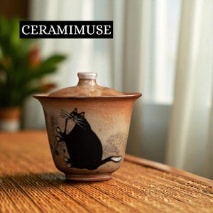 Pode incluir: Uma chávena de chá de cerâmica com tampa, com uma ilustração de um gato preto. A chávena tem um design gradiente castanho e bege, com o texto "CERAMIMUSE" num retângulo preto. A chávena está numa superfície de madeira.