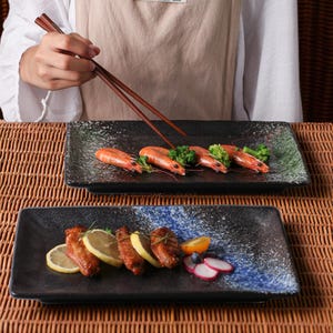 May include: Two rectangular black ceramic plates with food. One plate has shrimp and broccoli, the other has chicken wings, lemon slices, and vegetables. A person is holding chopsticks over the top plate.