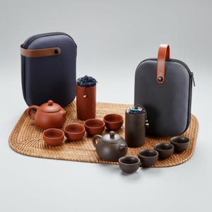 Könnte beinhalten: Ein Teeservice mit zwei Teekannen, mehreren kleinen Tassen und zwei Tragetaschen. Die Teekannen und Tassen sind braun, während die Koffer dunkelblau und grau mit Ledergriffen sind. Das Set ist auf einer gewebten Matte angeordnet.