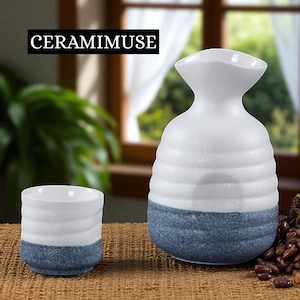 May include: A white and blue ceramic sake set, including a bottle and a cup. The bottle has a unique shape with a wide base and a narrow neck. The cup is cylindrical. The set is on a brown surface. The text "CERAMIMUSE" is in a black rectangle.