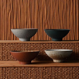 May include: Four ceramic bowls in varying colors: gray, dark teal, reddish-brown, and speckled beige. Each bowl has a wide, shallow design with a slightly textured surface. The bowls are displayed on a wooden shelf against a textured background.