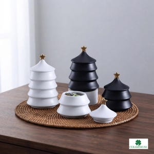 May include: A set of ceramic containers shaped like tiered Christmas trees with matte white and black finishes. Each piece features a small gold-coloured star topper. One white container is shown open, revealing its interior, while others have lids.