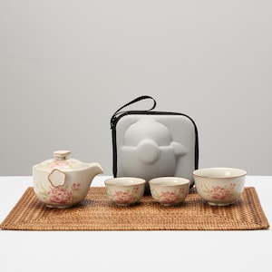 May include: A cream-coloured tea set with pink floral designs, including a teapot, three teacups, and a carrying case. The set is arranged on a woven mat, ideal for tea enthusiasts.