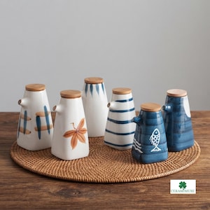 May include: A set of six ceramic oil dispensers with wooden lids, featuring various hand-painted designs including blue stripes, a brown leaf, a white fish, and blue cross-hatch patterns on white and blue glazed bodies with pouring spouts.