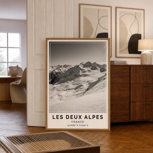 Puede incluir: Un cuadro vertical rectangular con marco de madera clara que muestra una fotografía en blanco y negro de picos montañosos nevados. El texto LES DEUX ALPES, FRANCE y las coordenadas 45.0069° N, 6.1245° E están impresos en negro en la parte inferior.