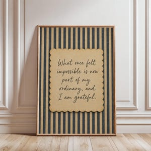 Può includere: Una stampa artistica incorniciata con un ritaglio di carta color marrone chiaro dai bordi smerlati, centrato su uno sfondo a righe verticali blu scuro e marrone chiaro. Il testo recita: What once felt impossible is now part of my ordinary, and I am grateful.