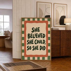 May include: A rectangular framed wall art print featuring a green and white checkered border surrounding a cream-coloured centre with a wavy orange frame. The text reads, "SHE BELIEVED SHE COULD, SO SHE DID" in dark green, bold, rounded lettering.