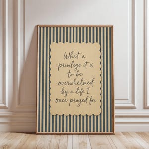 May include: A framed wall art print featuring a tan scalloped-edge card with the black text: What a privilege it is to be overwhelmed by a life I once prayed for. The background consists of vertical navy blue and tan stripes within a wooden frame.