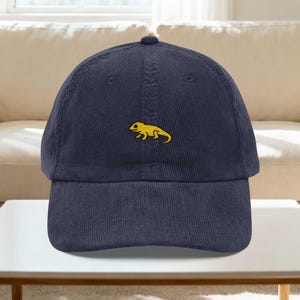 Könnte beinhalten: Marineblaue Cord-Baseballkappe mit einem kleinen, gestickten gelben Eidechsen-Design. Die Kappe hat einen gebogenen Schirm und einen verstellbaren Riemen. Ein lässiges Accessoire.