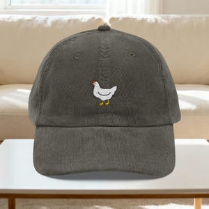 May include: A gray corduroy baseball cap featuring an embroidered white chicken with a red comb and yellow feet. The cap has a curved brim and a top button. The hat is displayed on a white table.