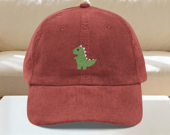 May include: A corduroy baseball cap in a deep red hue. The cap features a green dinosaur embroidered on the front. The dinosaur has white accents and a friendly expression. The cap has a curved brim and a rounded top.