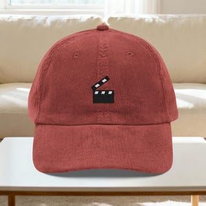 May include: A brick-red corduroy baseball cap featuring a black and white film clapperboard logo embroidered on the front. The cap has a curved brim and an adjustable strap. This hat is a stylish accessory for film enthusiasts.